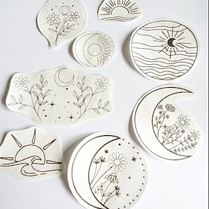 May include: A collection of cream-colored, die-cut stickers featuring black line art designs. The stickers depict celestial and floral themes, including moons, suns, flowers, and waves. The stickers vary in shape and size, with a stitched border.