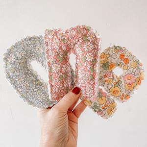 Peut inclure: Trois lettres en tissu floral dans une main. Les lettres ont la forme des lettres "O", "M" et "O". Le tissu a un fond blanc avec de petites fleurs dans les tons de rose, jaune et bleu.