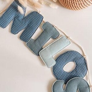 May include: A blue fabric banner spelling "MIO" with a grid pattern, strung on a natural cord. The letters are in varying shades of blue and are stuffed. A woven basket and dried flowers are in the background.