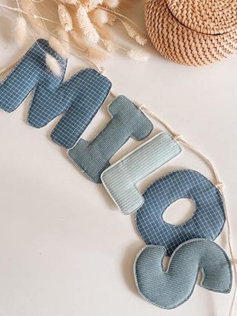 May include: A blue fabric banner spelling "MIO" with a grid pattern, strung on a natural cord. The letters are in varying shades of blue and are stuffed. A woven basket and dried flowers are in the background.