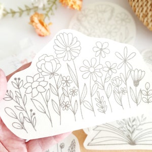 May include: A white sticker with a silver outline of a floral design. The design features a variety of flowers and leaves, including daisies, roses, and ferns.