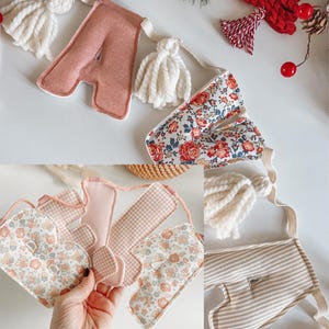 May include: A handmade banner with fabric letters, tassels, and decorative elements. The letters are in various patterns, including floral and striped designs. The color palette includes pink, white, and beige, with a festive holiday theme.