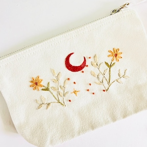 Pode incluir: Uma bolsa de lona branca com zíper com flores, folhas, uma lua crescente e estrelas bordadas. O bordado é em tons de vermelho, amarelo e verde.