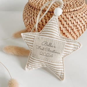 May include: A beige and white striped star-shaped ornament with the text "Bella's First Christmas 2024" embroidered on it.