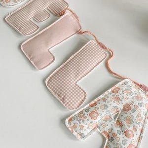 May include: A decorative banner spelling out the word "ONE". The letters are made of fabric in various patterns, including gingham and floral prints. The banner is strung together with a pink cord.