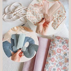 May include: A collection of handmade fabric decorations and materials. The image features a floral heart, gingham shapes, and the letters "MOM" in various pastel fabrics. Rolls of pink and floral fabric are also visible, along with a white rope.