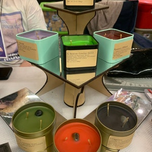 May include: A tiered display stand with six candles in various colours and scents. The candles are in metal tins with labels that include the names "Blackstede", "House", "D. Fenton (inviso bill)", "Mystery (Shack)", "Gimli", and "Afsorn".