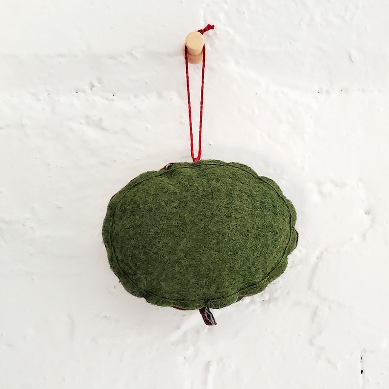 May include: A green felt ornament with a red string hanging from a wooden peg. The ornament is shaped like a circle and has a slightly textured surface.
