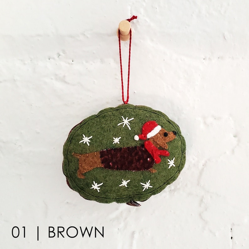 May include: Green felt Christmas ornament shaped like an oval with a brown dachshund wearing a red Santa hat and scarf. The ornament is decorated with white snowflakes.