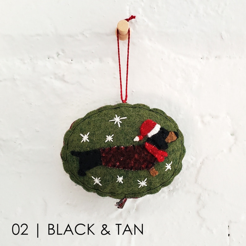 May include: A felt Christmas ornament shaped like a dachshund wearing a red Santa hat and scarf. The ornament is green with white snowflakes and the text "02 | BLACK & TAN".