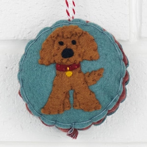 May include: A round felt ornament featuring a brown curly-haired dog with a red collar and a small yellow tag. The dog is centered on a teal circular background with a scalloped edge, and a red and white striped string is attached to the top.