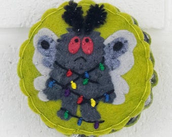May include: A round, lime green felt ornament featuring a dark gray moth character with large red eyes, black antennae, and light gray wings. The moth is wrapped in a string of colorful miniature Christmas lights in red, blue, yellow, and green.