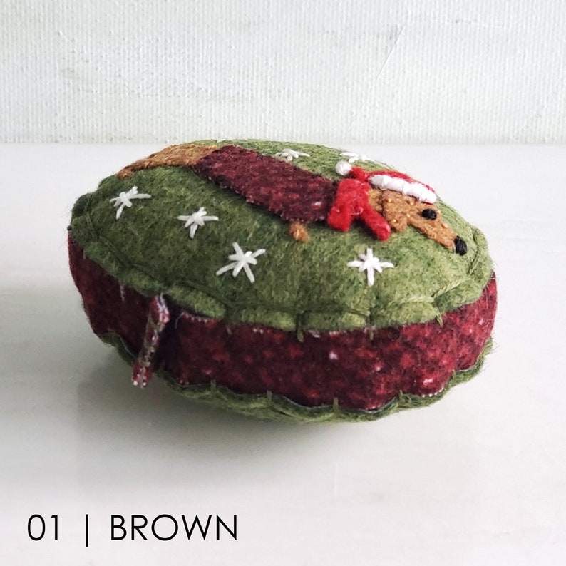 May include: A felt Christmas ornament shaped like a dachshund wearing a Santa hat. The ornament is green with white snowflakes and a red and brown trim.