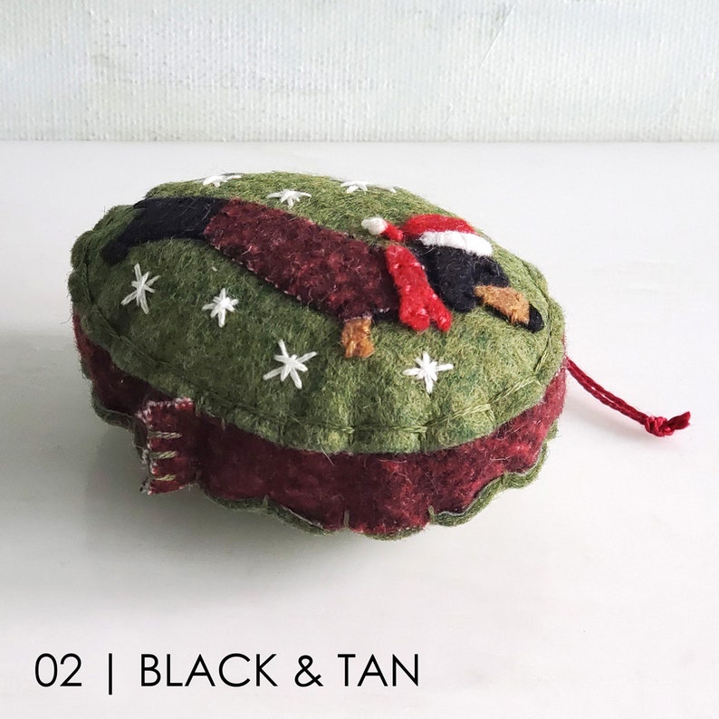 May include: Felt Christmas ornament shaped like a dachshund wearing a Santa hat. The ornament is green with white snowflakes and a red and burgundy trim. The dachshund is black and tan.