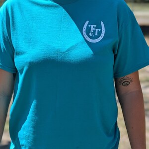 May include: Teal-colored t-shirt with a white horseshoe logo on the upper left chest. The logo features the letters "TT" and the number "4L" inside the horseshoe. The shirt has short sleeves and a crew neck.