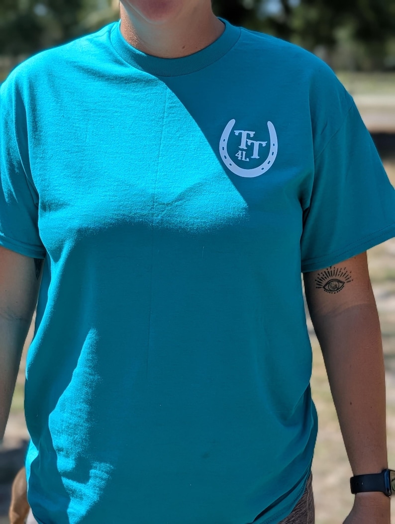 May include: Teal-colored t-shirt with a white horseshoe logo on the upper left chest. The logo features the letters "TT" and the number "4L" inside the horseshoe. The shirt has short sleeves and a crew neck.