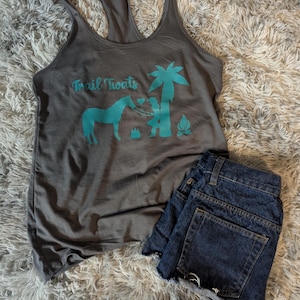 May include: Gray tank top with a teal design of a horse, a person, a palm tree, and a campfire. The text "Trail Treats" is printed on the front of the shirt.