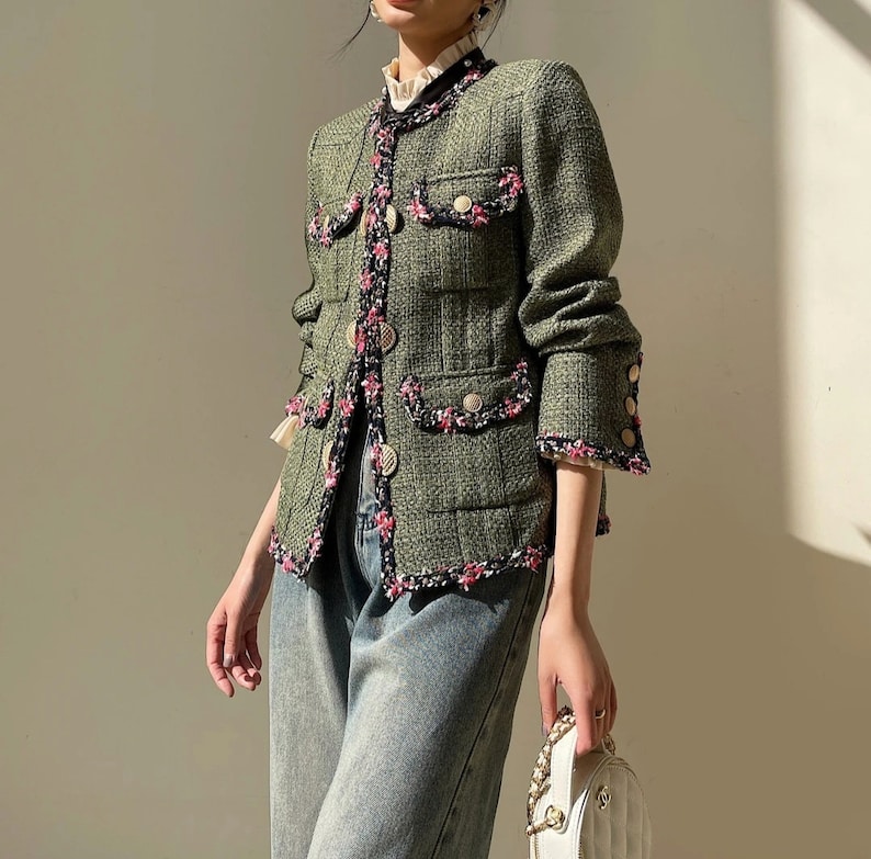 Op de afbeelding: Een groen tweed jasje met roze en witte bies. Het jasje heeft een knoopsluiting en klepzakken.