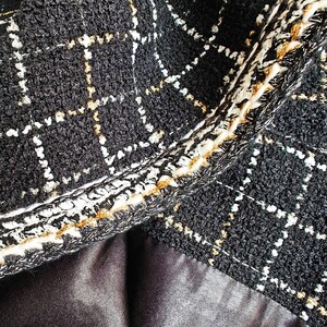 May include: Close-up of black tweed fabric with a gold and white geometric pattern. The fabric has a textured, woven look and a contrasting trim.