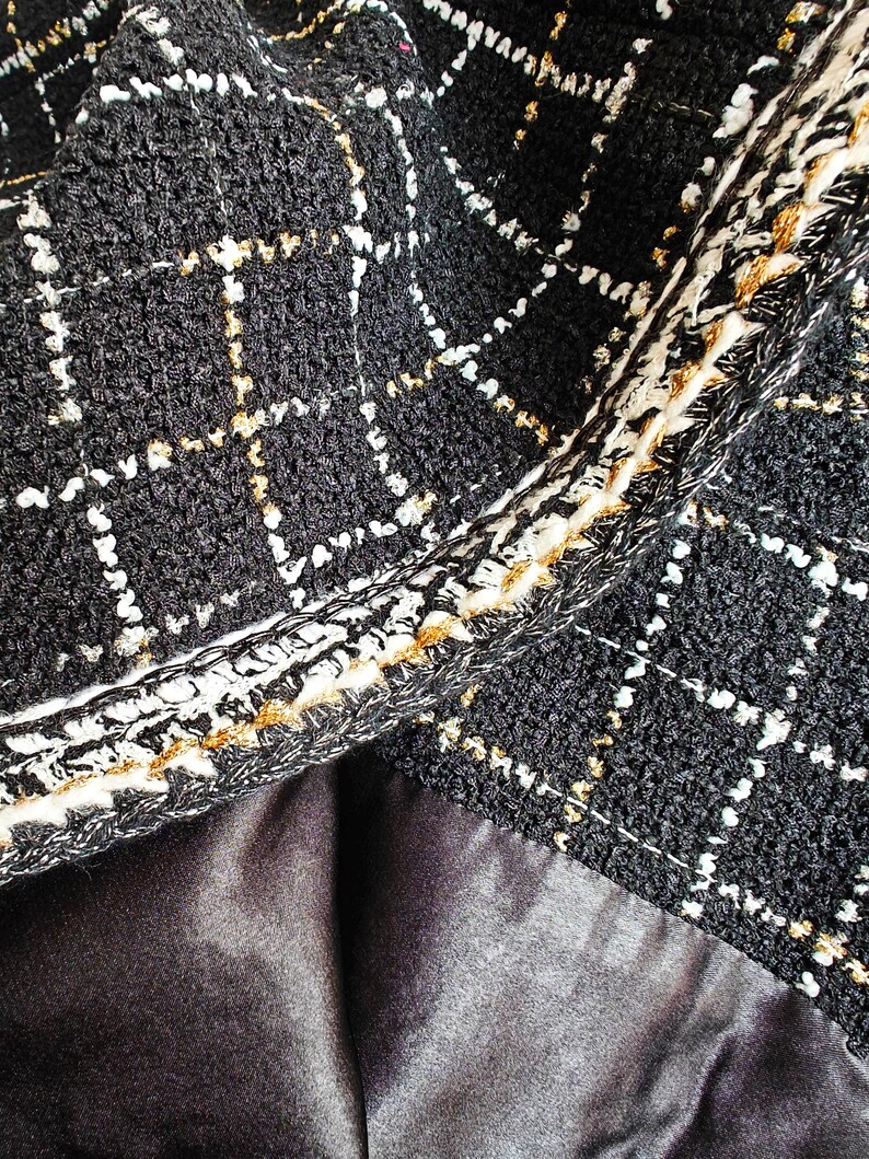 May include: Close-up of black tweed fabric with a gold and white geometric pattern. The fabric has a textured, woven look and a contrasting trim.