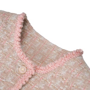 K&ouml;nnte beinhalten: Nahaufnahme einer hellrosa und beigefarbenen Tweedjacke mit dekorativer rosa Bord&uuml;re um Kragen und Vorderseite. Die Jacke hat einen einzelnen, runden, perlenartigen Knopf. Der Stoff hat einen dezenten Schimmer.