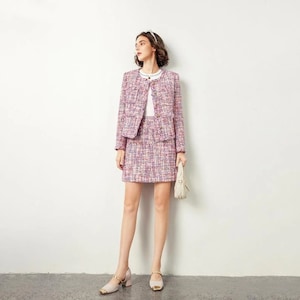 Puede incluir: Un traje de falda de tweed rosa y blanco con una falda corta y una chaqueta a juego. La chaqueta tiene un escote redondo y cierre de botón. La falda tiene una cintura alta y una silueta recta.