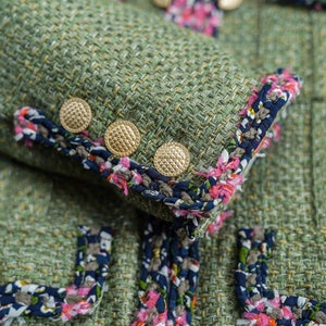 Op de afbeelding: Close-up van een groen tweed jasje met een decoratieve rand van roze, blauwe en witte bloemen. Het jasje heeft goudkleurige knopen.