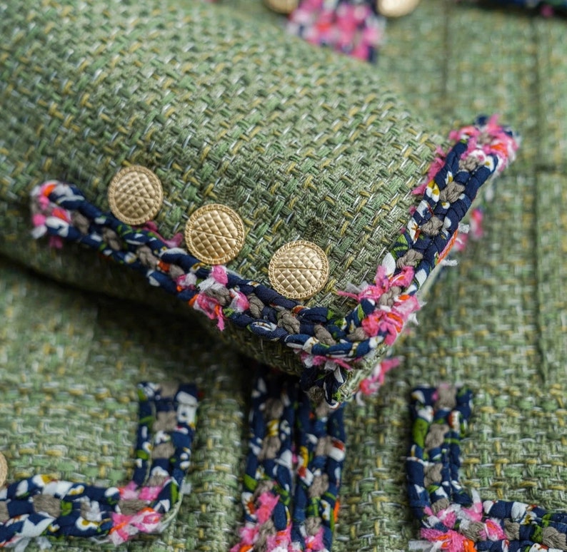 Op de afbeelding: Close-up van een groen tweed jasje met een decoratieve rand van roze, blauwe en witte bloemen. Het jasje heeft goudkleurige knopen.