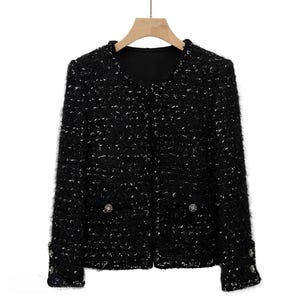 May include: A black tweed jacket with silver flecks, featuring a round neckline and two front pockets. The jacket has decorative buttons on the front and cuffs, and is displayed on a wooden hanger.
