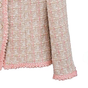 K&ouml;nnte beinhalten: Nahaufnahme einer hellrosa und cremefarbenen Tweedjacke. Die Jacke hat ein strukturiertes Gewebe mit rosa Akzenten und eine dekorative rosa Bord&uuml;re an den Kanten und Manschetten. Zwei beige Kn&ouml;pfe sind sichtbar.