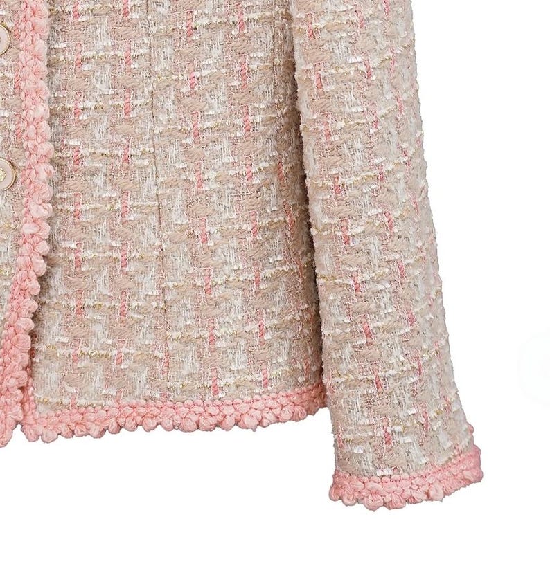 K&ouml;nnte beinhalten: Nahaufnahme einer hellrosa und cremefarbenen Tweedjacke. Die Jacke hat ein strukturiertes Gewebe mit rosa Akzenten und eine dekorative rosa Bord&uuml;re an den Kanten und Manschetten. Zwei beige Kn&ouml;pfe sind sichtbar.