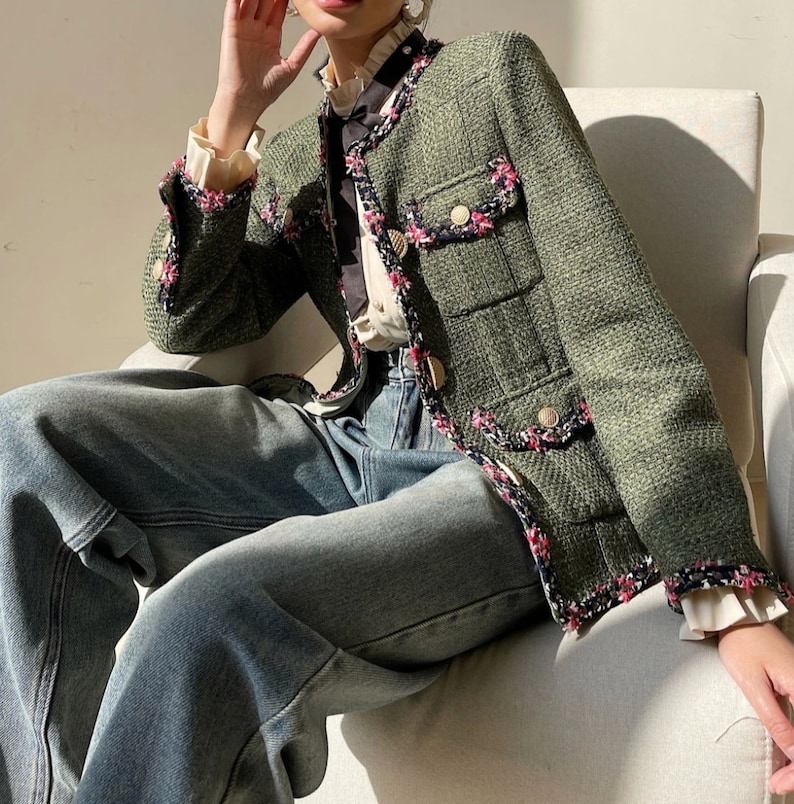 Op de afbeelding: Een groen tweed jasje met roze en zwarte bies, met een gouden knoopsluiting en een grote klepzak. Het jasje wordt gedragen over een wit blouse met een zwarte strik en een blauwe jeans.