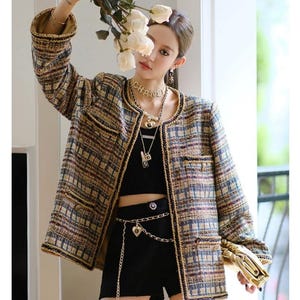 May include: A brown, blue, and yellow tweed jacket with a black trim and a gold chain belt. The jacket has a round neckline and long sleeves. The model is wearing a black top and black shorts.