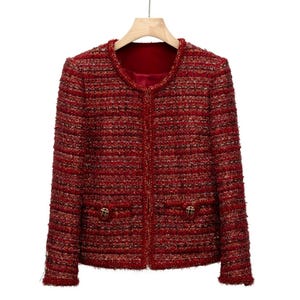 Puede incluir: Una chaqueta de tweed roja con botones dorados y bolsillos. La chaqueta tiene un cuello redondo y una cremallera.