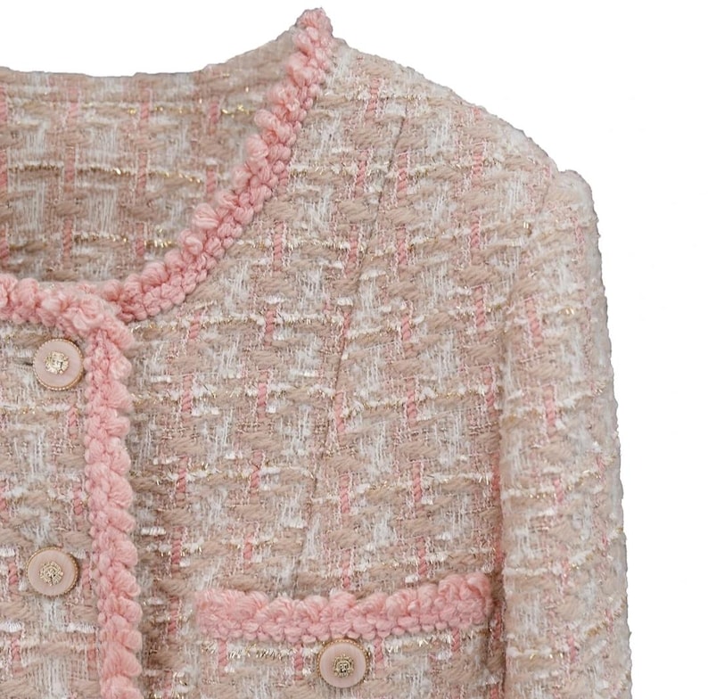 K&ouml;nnte beinhalten: Nahaufnahme einer rosa-beigen Tweedjacke. Die Jacke hat einen runden Ausschnitt, dekorative Kn&ouml;pfe und rosa Bes&auml;tze. Der Stoff hat eine gewebte Textur mit dezenten Goldf&auml;den.