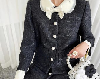 May include: A black tweed jacket with a white lace collar and a large white bow. The jacket has pearl buttons and a pocket on the front. The model is also wearing a black skirt and a white pearl necklace.