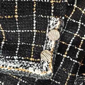 May include: Close-up of a black and white tweed fabric with gold thread accents. Two silver buttons are visible on the fabric.