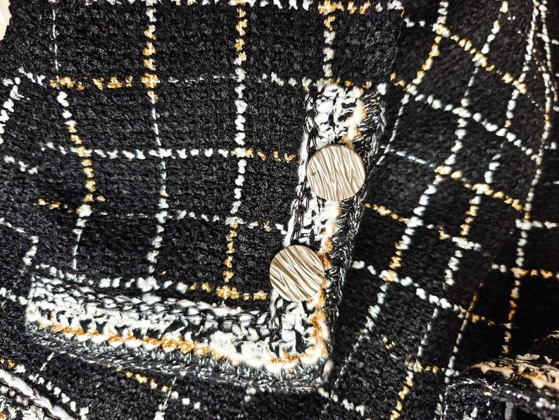 May include: Close-up of a black and white tweed fabric with gold thread accents. Two silver buttons are visible on the fabric.