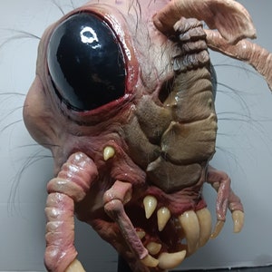 Op de afbeelding: Close-up van een gedetailleerd, roze en bruin alien-wezensmasker met een groot zwart oog, scherpe tanden en een textuur.