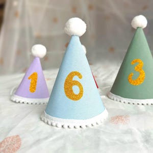 May include: Three cone-shaped party hats in light purple, light blue, and sage green. Each hat features a white pom-pom on top, a white pom-pom trim along the bottom edge, and a large glittery gold number on the front: 1, 6, and 3 respectively.
