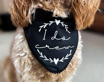 May include: A black triangular pet bandana featuring a white graphic of a circular wreath with hearts at the top and bottom. The text "I do crew" is printed in white script in the center. The edges of the bandana are finished with small black pom-poms.