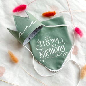 May include: A sage green fabric crown with the word one in white script and a matching sage green triangular bandana featuring the white text IT'S MY BIRTHDAY with a small star icon and white pom-pom trim along the bottom edge.