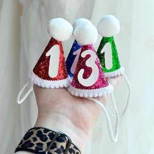 May include: Four miniature cone-shaped party hats covered in red, blue, magenta, and green glitter. Each hat features a white pom-pom on top, a white pom-pom trim at the base, a white elastic chin strap, and a large white number one or three.