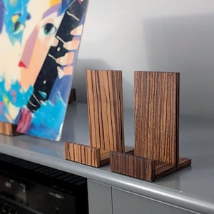 May include: Two L-shaped wooden display stands featuring a distinct dark brown and tan striped wood grain pattern. Each stand consists of a vertical back panel and a horizontal base with a small front lip, designed to hold items upright.