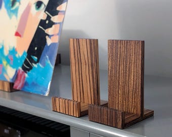 May include: Two L-shaped wooden display stands featuring a distinct dark brown and tan striped wood grain pattern. Each stand consists of a vertical back panel and a horizontal base with a small front lip, designed to hold items upright.