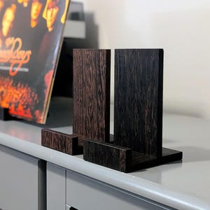 Pode incluir: Suporte de disco de madeira com acabamento marrom escuro e preto. O suporte tem dois suportes verticais e uma base, projetados para exibir discos de vinil. Um álbum de disco é visível no fundo.