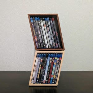 May include: Angled wooden DVD and Blu-ray storage unit. The top section is dark brown, and the bottom is light brown. The unit holds various movie titles, including "Sherlock Holmes," "Robocop," and "Aquaman."