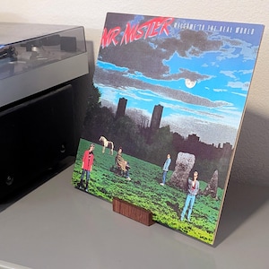 A vinyl record album cover for Mr. Mister's "Welcome to the Real World" album. The cover art features a surreal landscape with figures, a horse, and the band's name in red. The album is displayed on a wooden stand.