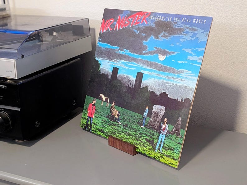 A vinyl record album cover for Mr. Mister's "Welcome to the Real World" album. The cover art features a surreal landscape with figures, a horse, and the band's name in red. The album is displayed on a wooden stand.