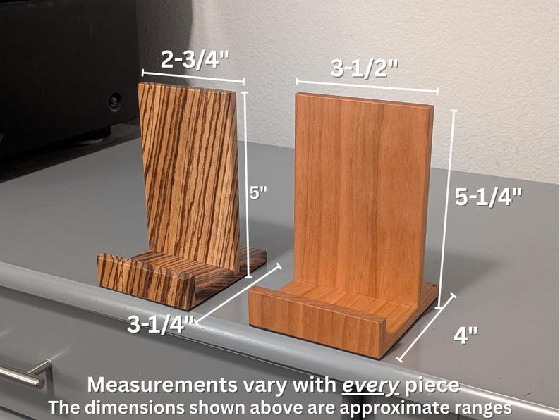 Two wooden record display stands shown side by side on a gray surface, one made of zebrawood and the other of cherry wood. White measurement lines indicate approximate dimensions: the zebrawood stand measures about 5 inches tall, 3-1/4 inches deep, and 2-3/4 inches wide, while the cherry stand measures about 5-1/4 inches tall, 4 inches deep, and 3-1/2 inches wide. Text below notes that measurements vary with every handmade piece.